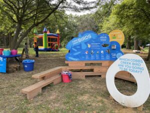 A Playful Learning structure, including wooden bench seating, a sign with illustrations of local birds, and a photo opportunity shaped like an egg which says "Which Cobbs Creek Bird Are You Going to Be Today?"
