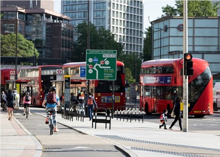 A city street including a huge protected bike lane, lots of bike racks, many cyclists and pedestrians, and large buses.